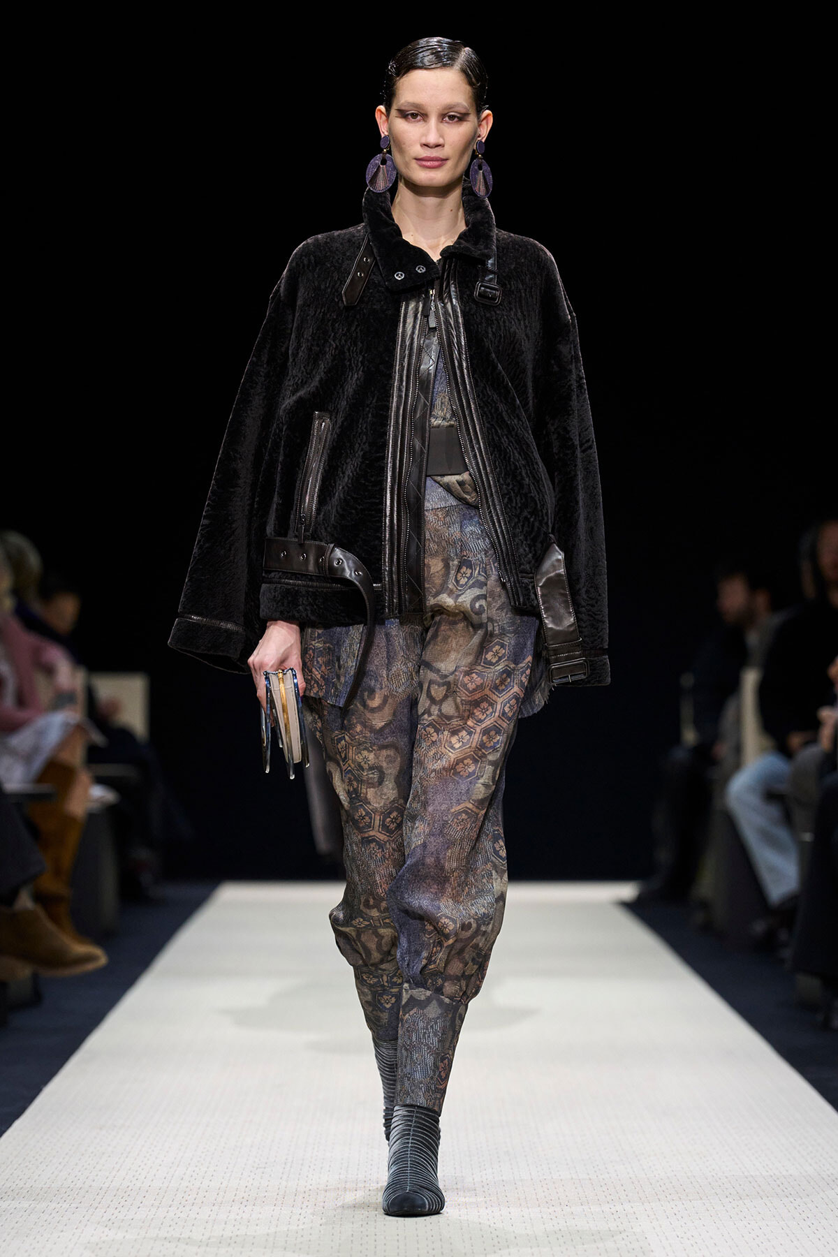 Model walking a runway in a black fur coat, patterned pants, and striped heeled boots, holding a small clutch.