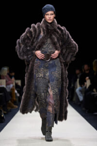 Model walking a fashion runway in a dark fur coat over a patterned dress with a wide belt and gray boots, head wearing a blue knit cap.