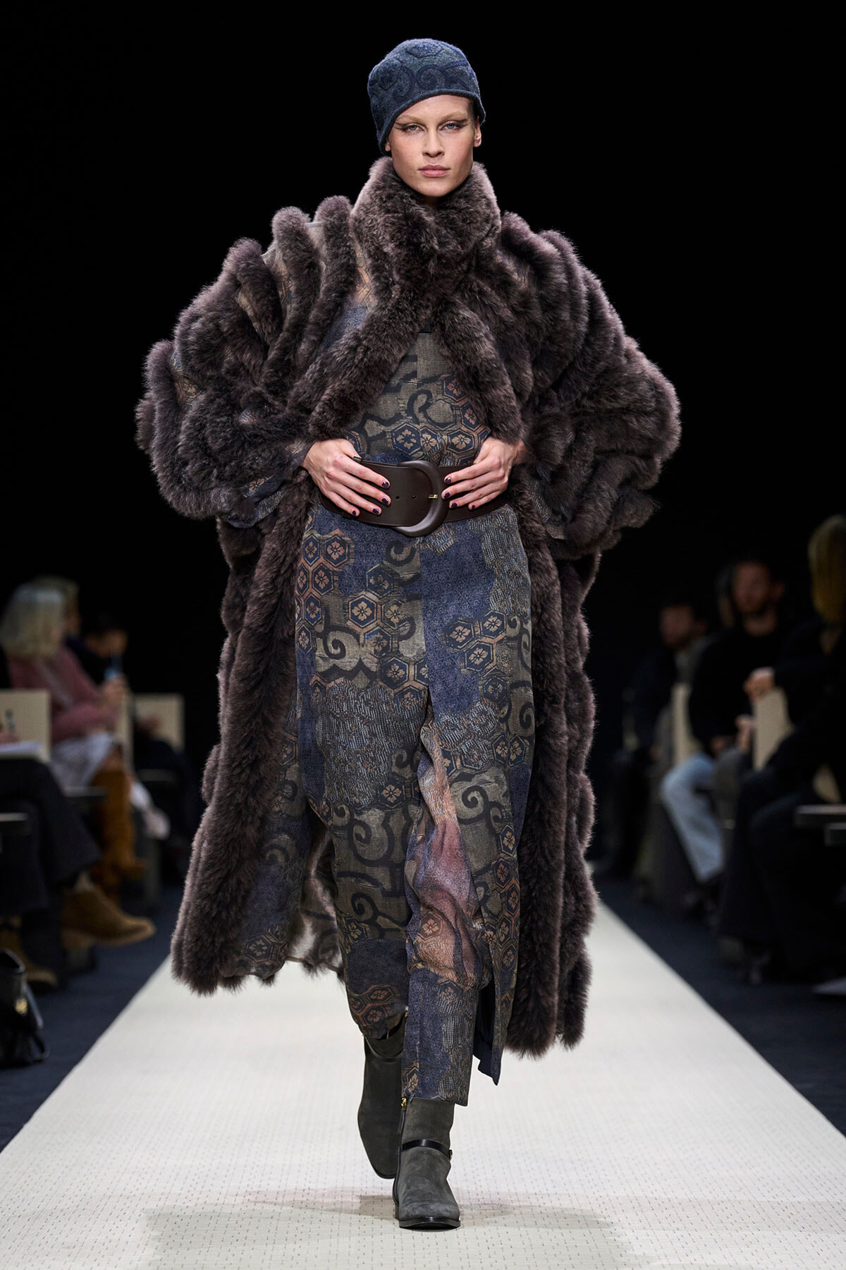 Model walking a fashion runway in a dark fur coat over a patterned dress with a wide belt and gray boots, head wearing a blue knit cap.