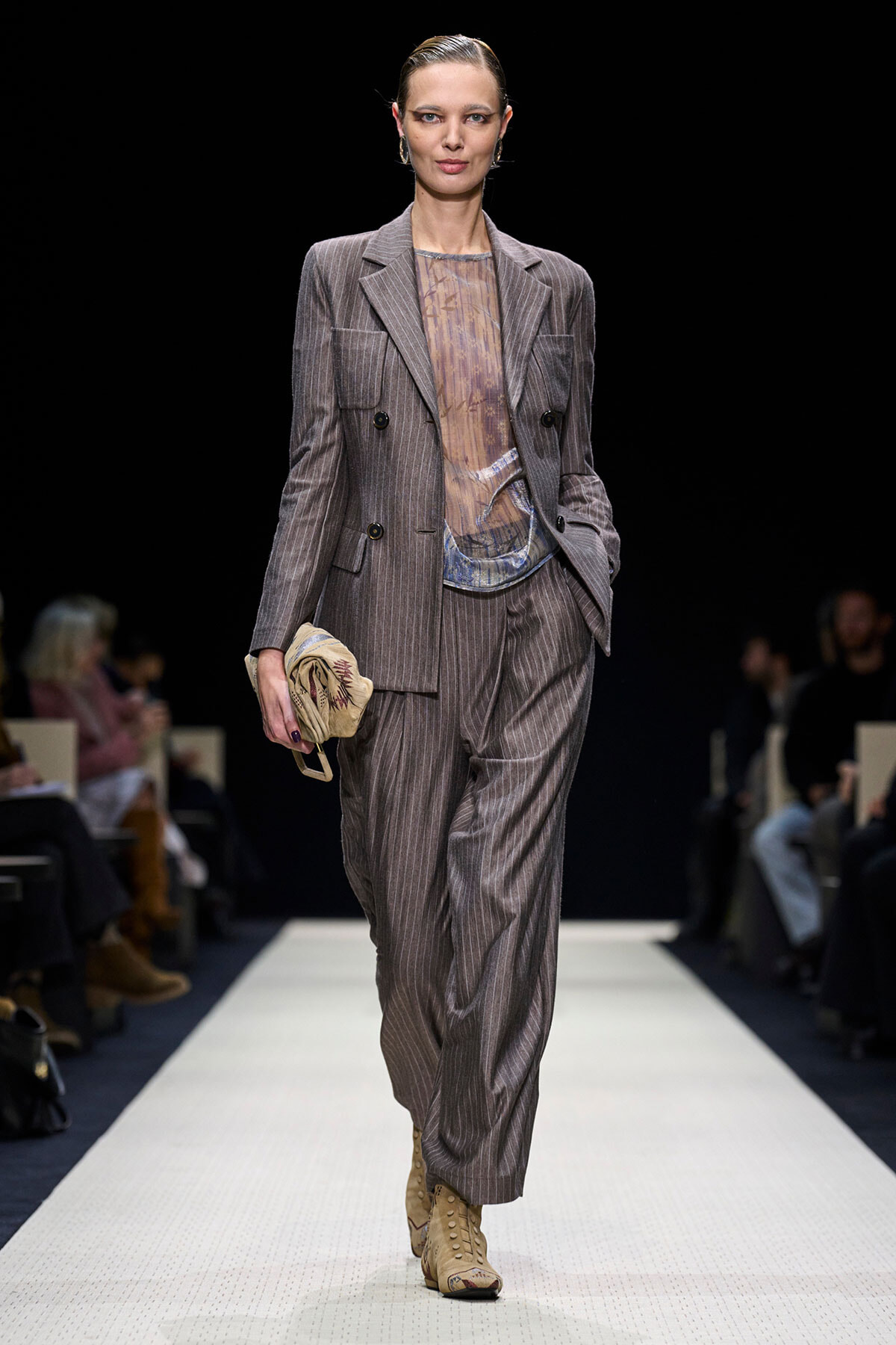 Runway model wearing a gray pinstripe suit with a sheer top, beige boots, and a beige clutch in hand.