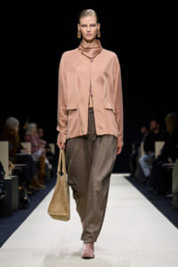 Model walking a runway in a pink satin blouse with a high neck and matching pale pink shoes, carrying a tan tote bag.