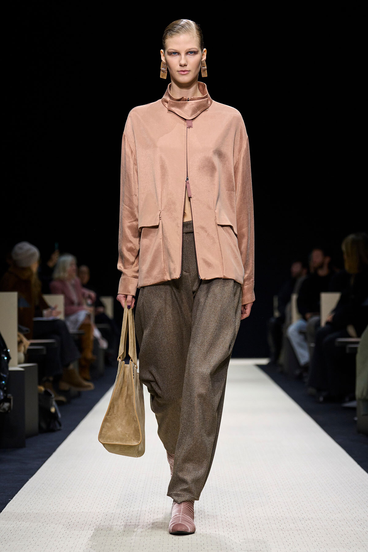Model walking a runway in a pink satin blouse with a high neck and matching pale pink shoes, carrying a tan tote bag.
