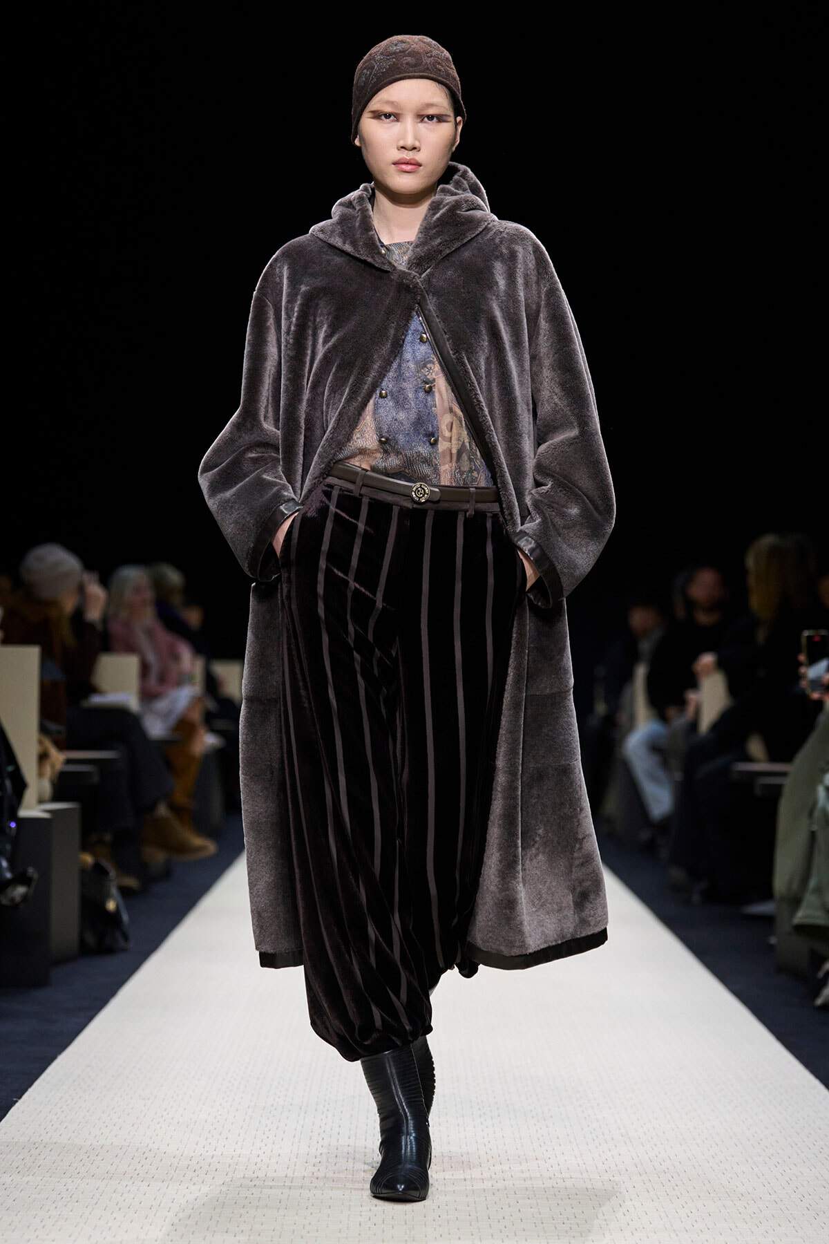 Model walks the runway in a dark velvet oversized coat over a patterned blouse and striped velvet pants, paired with black ankle boots.