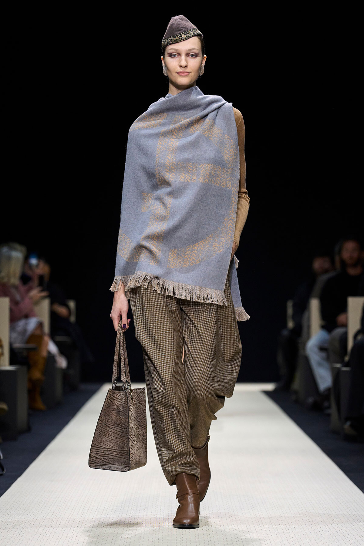 Model walking a runway in a blue fringed shawl with gold patterns over a tan top, loose brown trousers, brown boots, and a textured brown handbag.