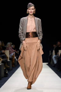 Model walks the runway in a peach satin maxi dress with a draped skirt, paired with a gray blazer and wide metallic belt.