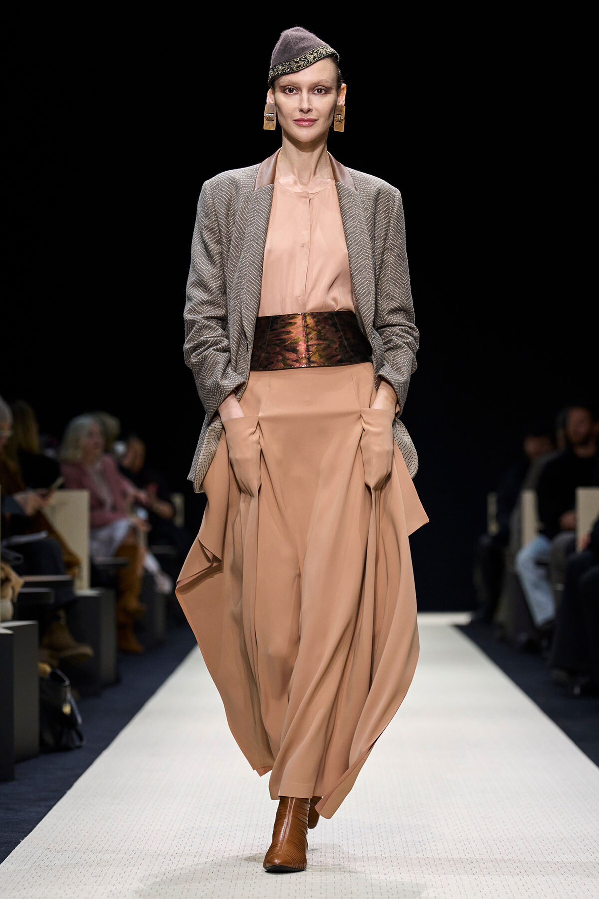 Model walks the runway in a peach satin maxi dress with a draped skirt, paired with a gray blazer and wide metallic belt.