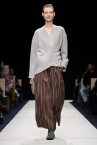 Runway model wearing a light gray wrap sweater with button accents and brown striped wide-leg pants, gray ankle boots, and a beige clutch.