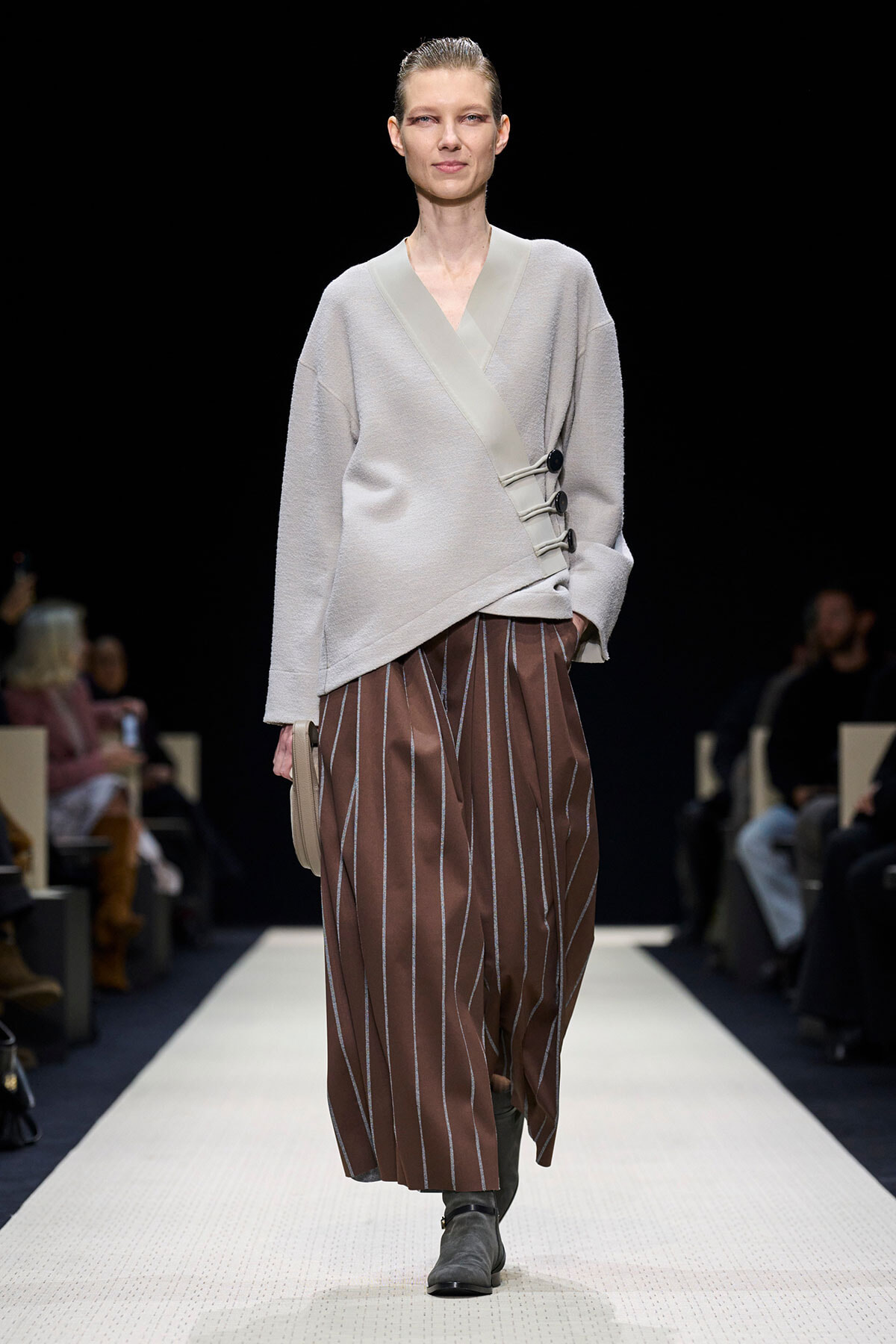 Runway model wearing a light gray wrap sweater with button accents and brown striped wide-leg pants, gray ankle boots, and a beige clutch.