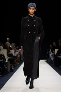 Model on a runway wearing a long black textured coat with gold buttons, blue patterned headscarf, and black ankle boots, holding a small handbag.