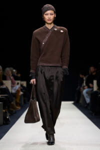 Model walking a fashion runway in a brown knit top with diagonal strap, wide dark trousers, and brown accessories; audience blurred in background.