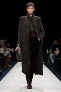 Model walks the runway in a long dark textured coat, matching cap, velvet brown pants, and stacked boots, carrying a gray clutch.