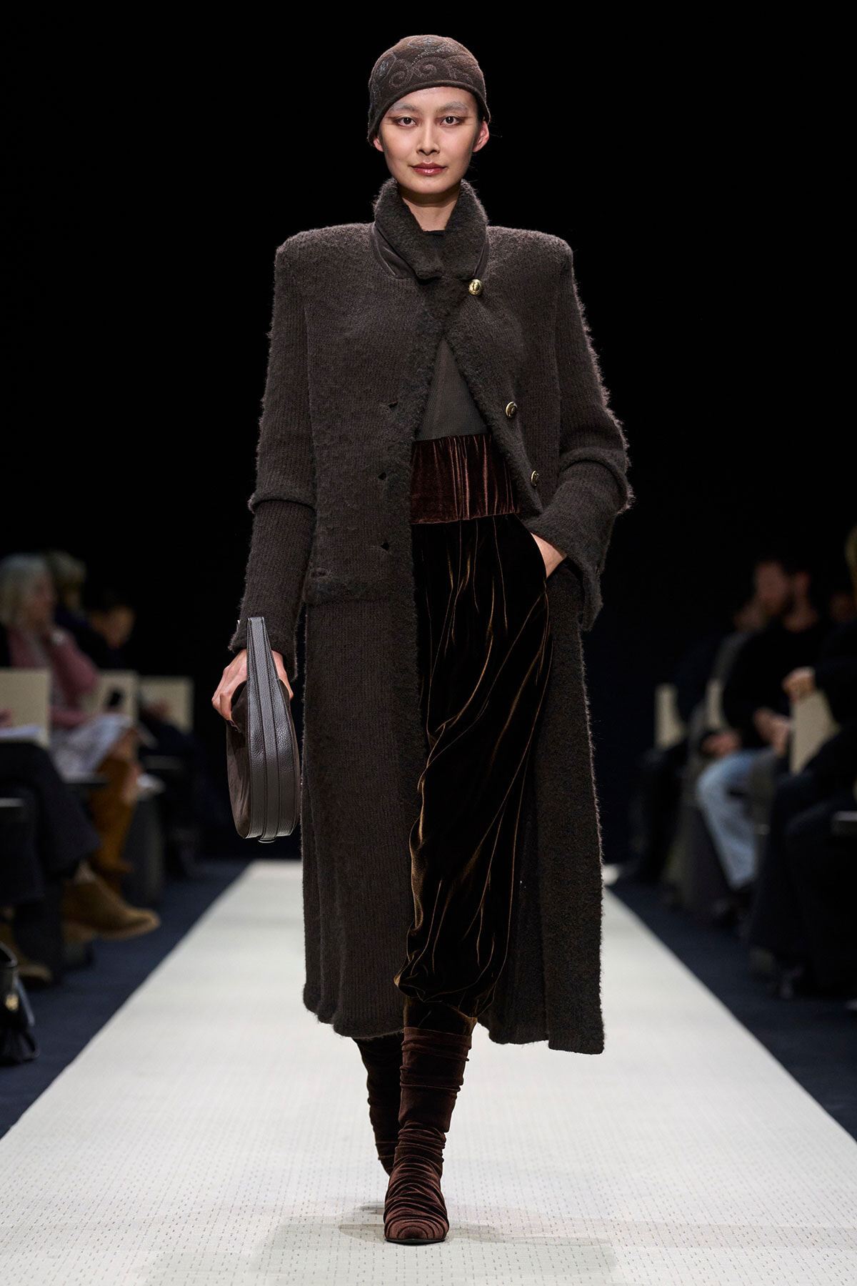 Model walks the runway in a long dark textured coat, matching cap, velvet brown pants, and stacked boots, carrying a gray clutch.