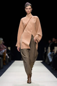 Model walks a runway in a loose peach blouse, brown textured trousers, and brown ankle boots; crossbody strap over her shoulder and a patterned headband.
