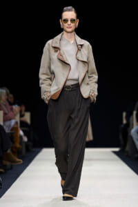 Model on a runway wearing a beige suede jacket with brown piping, gray sweater, and black wide-leg pants with a belt and round sunglasses.