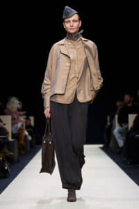 Model on a runway wearing a beige cropped jacket, loose black pleated pants, a patterned scarf, and a blue hat, carrying a dark tote bag.