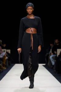 Model walking a runway in a long black coat-dress with a wide tan leather belt, headscarf, and black boots, holding a small black clutch against a dark backdrop.