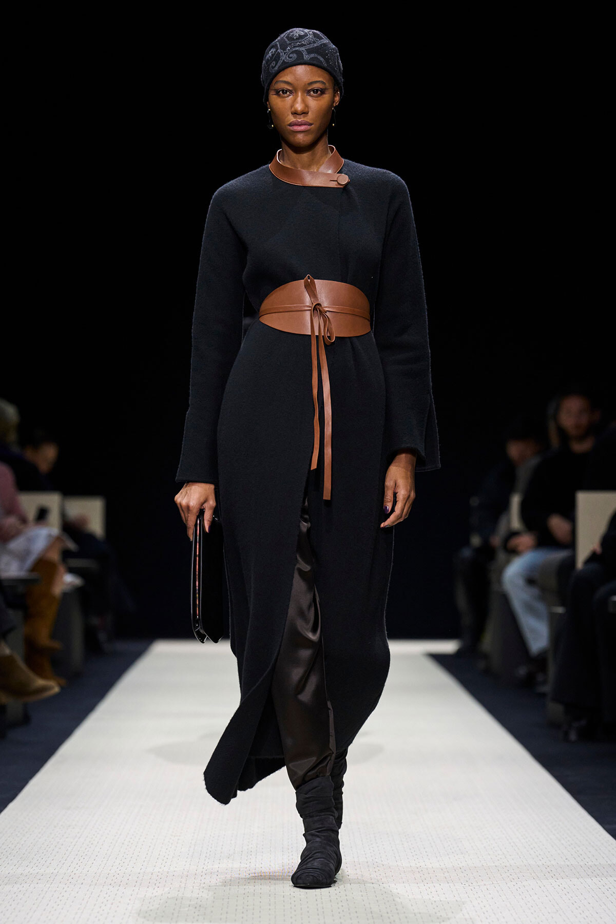 Model walking a runway in a long black coat-dress with a wide tan leather belt, headscarf, and black boots, holding a small black clutch against a dark backdrop.