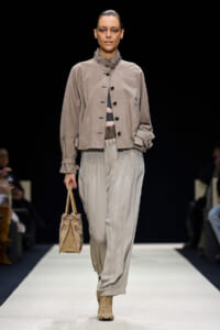 Runway model in a taupe cropped jacket with buttons, paired with wide beige pleated trousers, tan boots, and a small beige handbag.