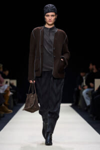Model walks a runway in a dark brown coat with light piping, gray top, and black patterned pants, carrying a brown suede bag.