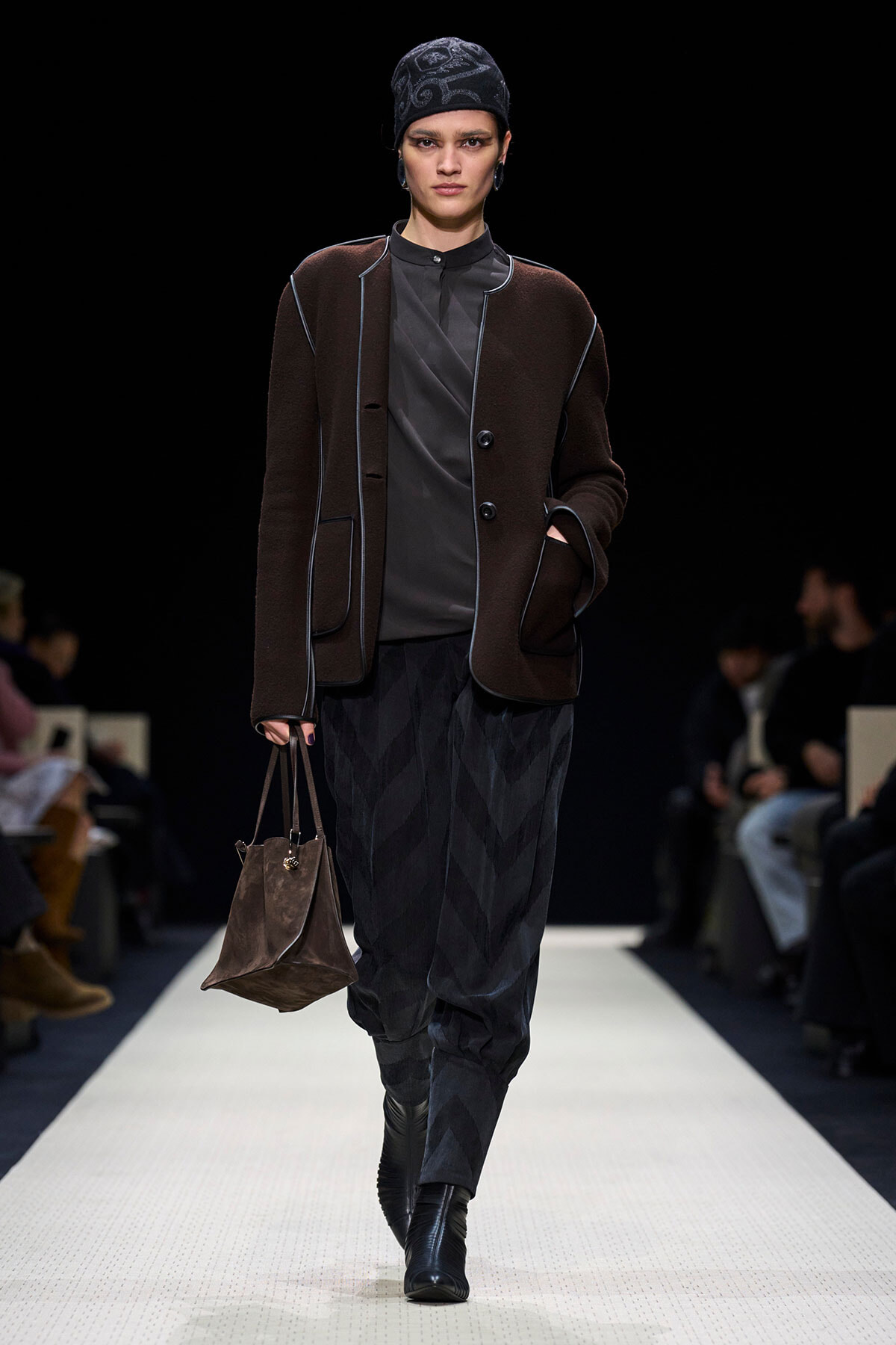 Model walks a runway in a dark brown coat with light piping, gray top, and black patterned pants, carrying a brown suede bag.