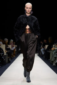 Model with platinum hair walks the runway in a black faux fur coat, metallic belt, and loose brown satin pants, gray sock boots visible.