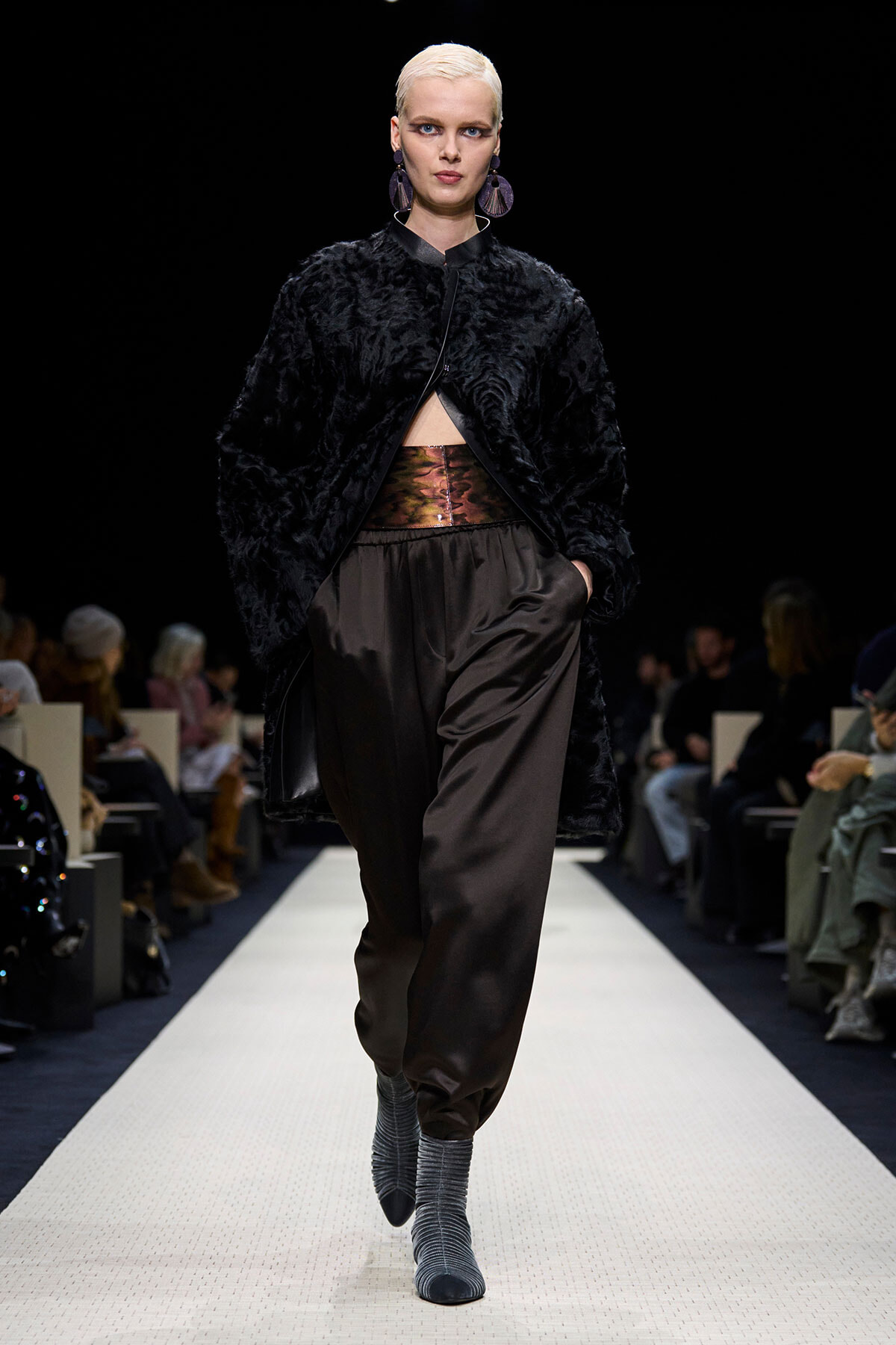 Model with platinum hair walks the runway in a black faux fur coat, metallic belt, and loose brown satin pants, gray sock boots visible.