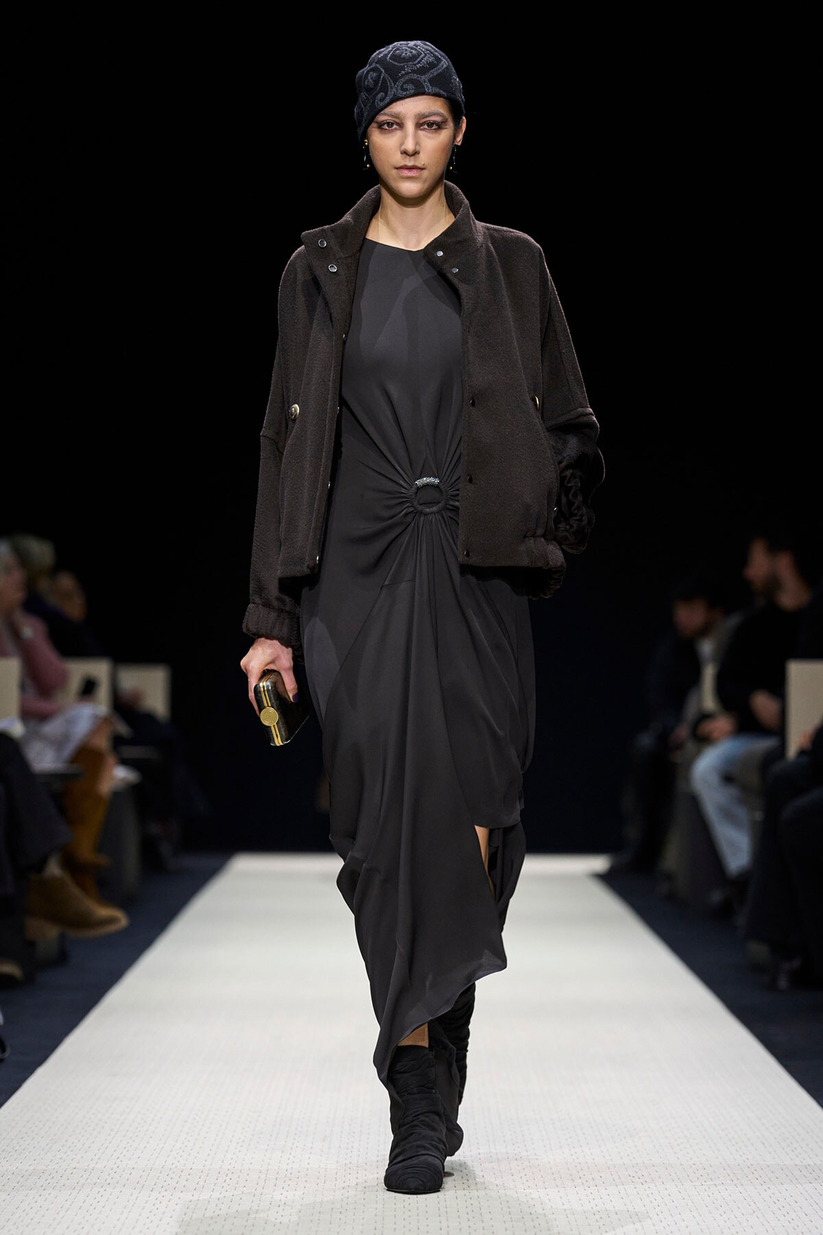 Female model on a runway wearing a dark, twisted-waist gray dress, a brown overcoat, and a patterned headscarf.