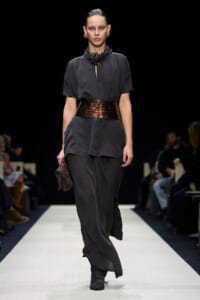 Model walks the runway in a dark grey tunic with a wide metallic belt, loose pants, black boots, and a chunky beaded necklace, holding a patterned clutch.