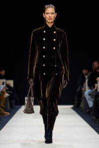 Model walking a dark runway in a long chocolate-brown velvet double-breasted coat with silver buttons, gloves, and a small patterned handbag.
