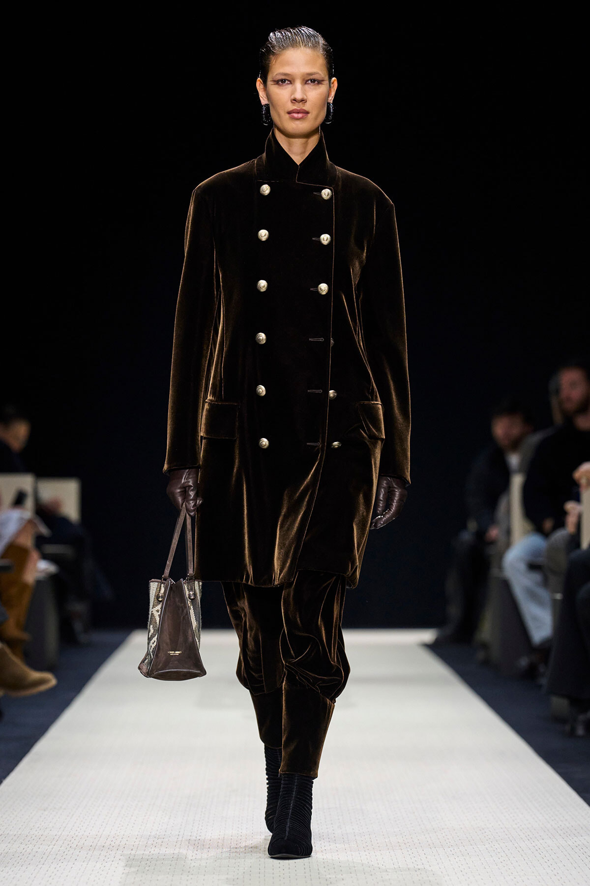 Model walking a dark runway in a long chocolate-brown velvet double-breasted coat with silver buttons, gloves, and a small patterned handbag.