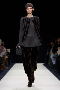 Model walks a runway in a dark velvet vest over a gray tunic, matching velvet pants, maroon boots, and a patterned headscarf.