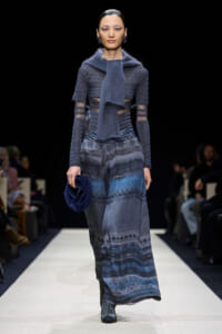 Model walking a runway in a blue-toned knit dress with a matching scarf and dark blue clutch.