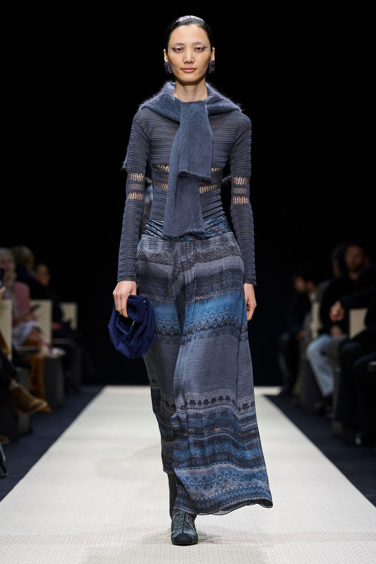 Model walking a runway in a blue-toned knit dress with a matching scarf and dark blue clutch.
