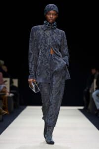 Model walking the runway in a textured blue-gray suit with a midriff cutout and matching boots, holding a small clutch.