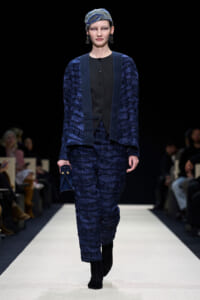 Model walking a fashion runway in a textured navy-blue suit with matching vest and headscarf, holding a small blue clutch.