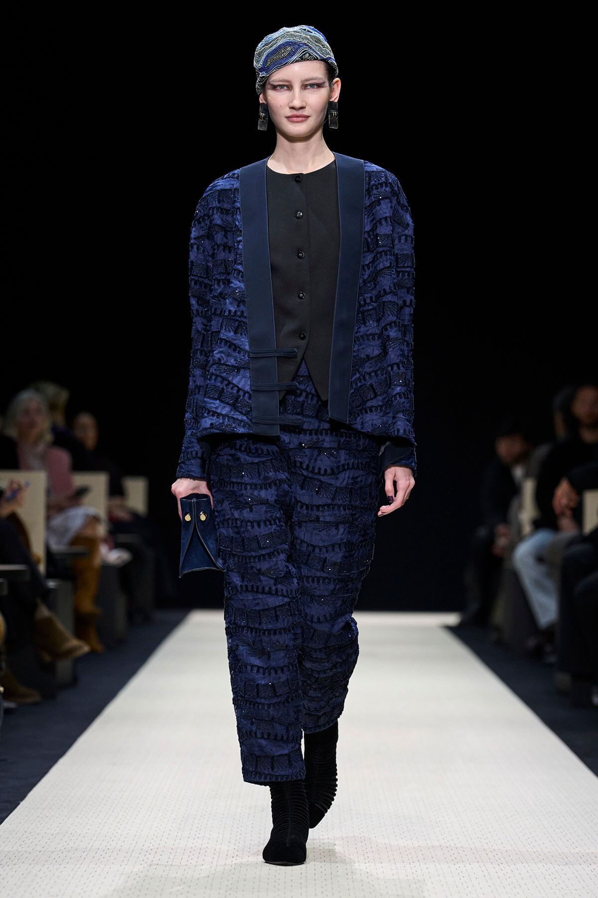 Model walking a fashion runway in a textured navy-blue suit with matching vest and headscarf, holding a small blue clutch.