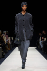 Model walking a fashion runway in a charcoal textured coat, gray trousers, and a patterned headscarf, carrying a small dark handbag.