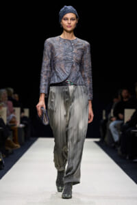 Model on a runway wearing a textured blue knit cardigan, loose gray pleated trousers, a blue headwrap, and gray boots, carrying a small clutch.