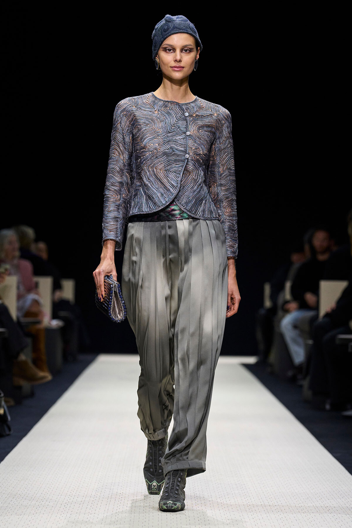 Model on a runway wearing a textured blue knit cardigan, loose gray pleated trousers, a blue headwrap, and gray boots, carrying a small clutch.