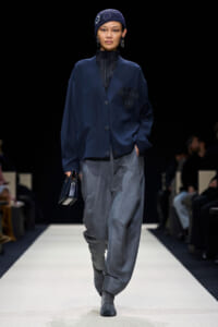 Model walking a runway in a navy oversized jacket with a patch detail and wide gray pants, wearing a knit cap and striped socks.