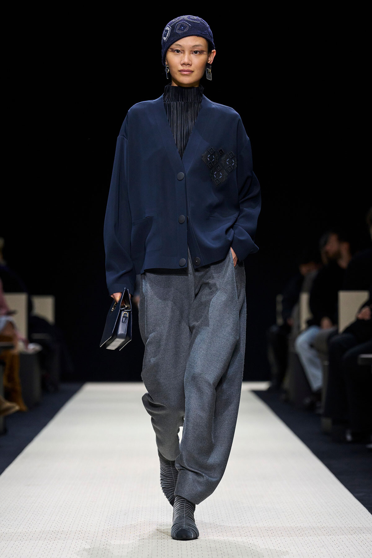 Model walking a runway in a navy oversized jacket with a patch detail and wide gray pants, wearing a knit cap and striped socks.