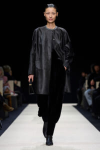 Model walking the runway in a dark oversized coat, black velvet pants, and ankle boots, holding a small black handbag.