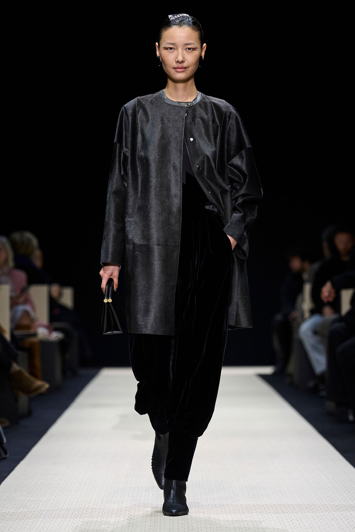 Model walking the runway in a dark oversized coat, black velvet pants, and ankle boots, holding a small black handbag.