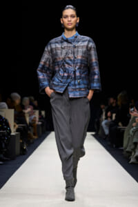 Model on a runway wearing a blue patterned double-breasted jacket with gray pleated trousers and gray ankle boots.