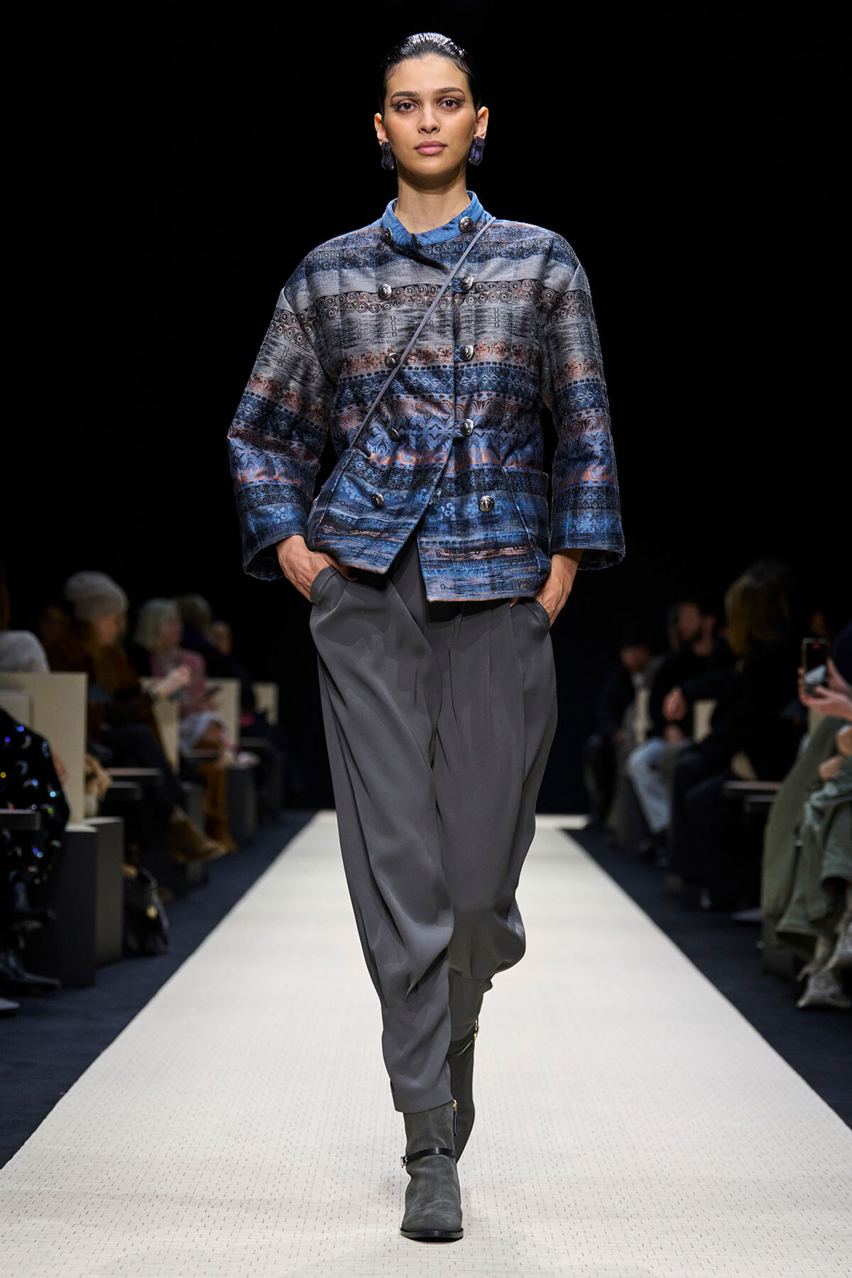 Model on a runway wearing a blue patterned double-breasted jacket with gray pleated trousers and gray ankle boots.