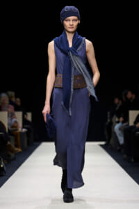 Model on a fashion runway wearing a blue layered dress, scarf, and matching knit hat with ankle boots.