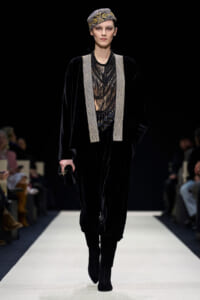 Model walking a runway in a black velvet outfit with silver-beaded trim and a jeweled headpiece on her head.