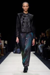 Model walks a fashion runway wearing a black velvet military-style jacket with silver buttons and pleated multicolored pants, carrying a small patterned handbag.