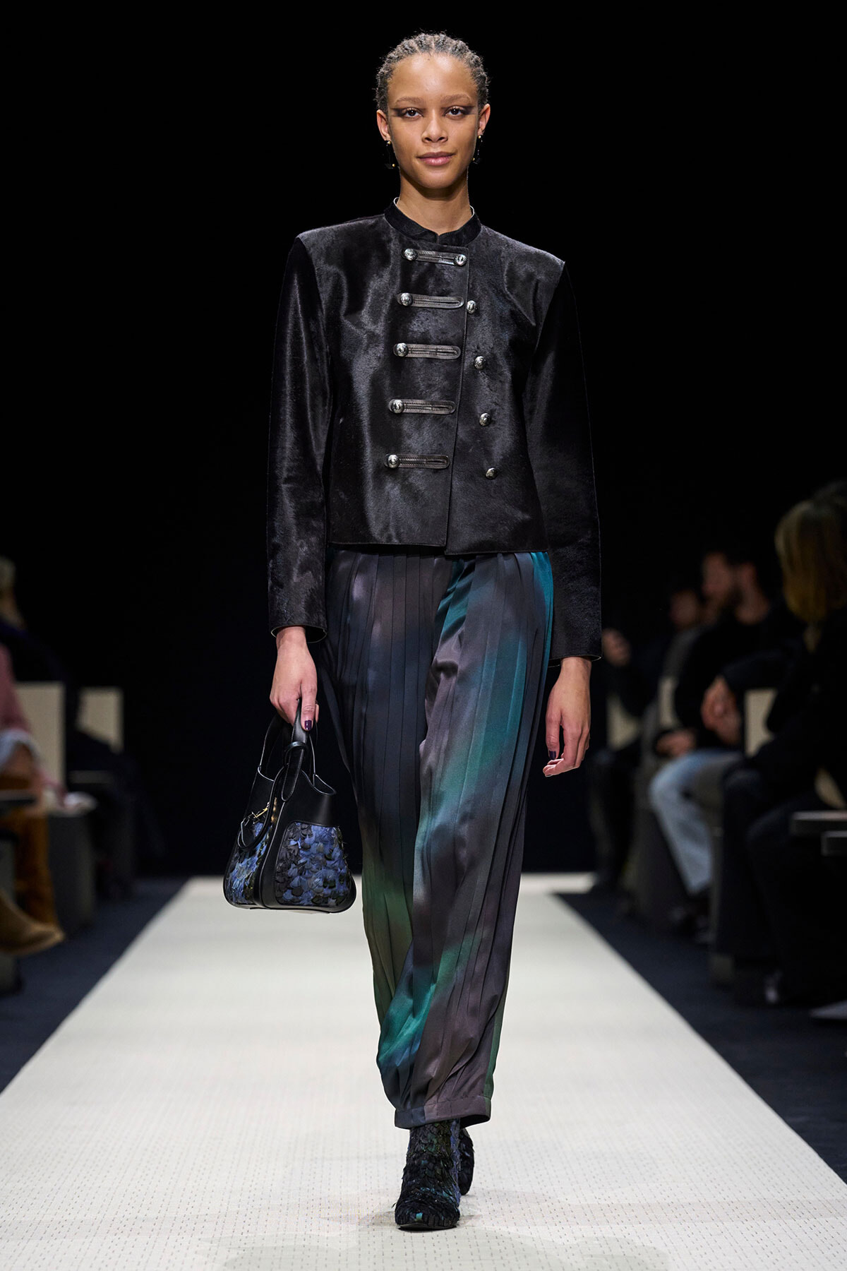 Model walks a fashion runway wearing a black velvet military-style jacket with silver buttons and pleated multicolored pants, carrying a small patterned handbag.