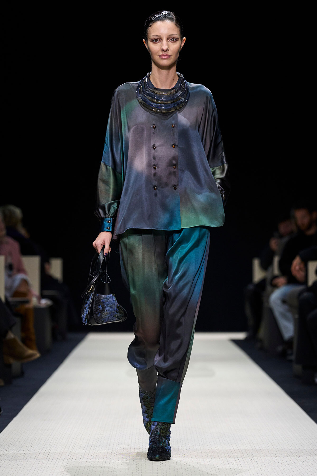 Model on a fashion runway wearing a loose satin gradient teal-to-gray blouse with matching pants, a chunky layered necklace, and teal boots, carrying a small patterned handbag.